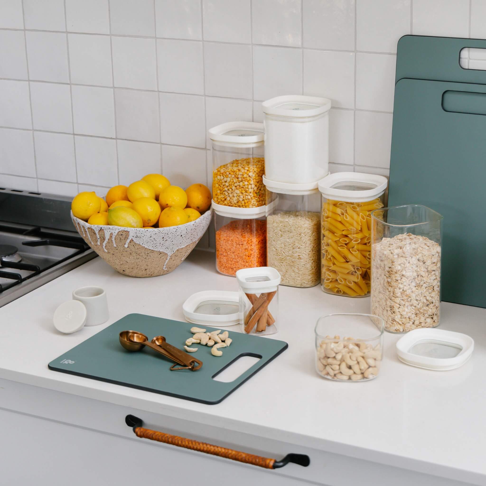 a white countertop with a green chopping board, glass food jars with different grains and nuts inside them and a set of measuring spoons