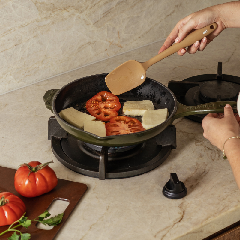 Praline Beige silicone round spatula stirring tomatoes and halloumi in a cast iron skillet on a gas stovetop.