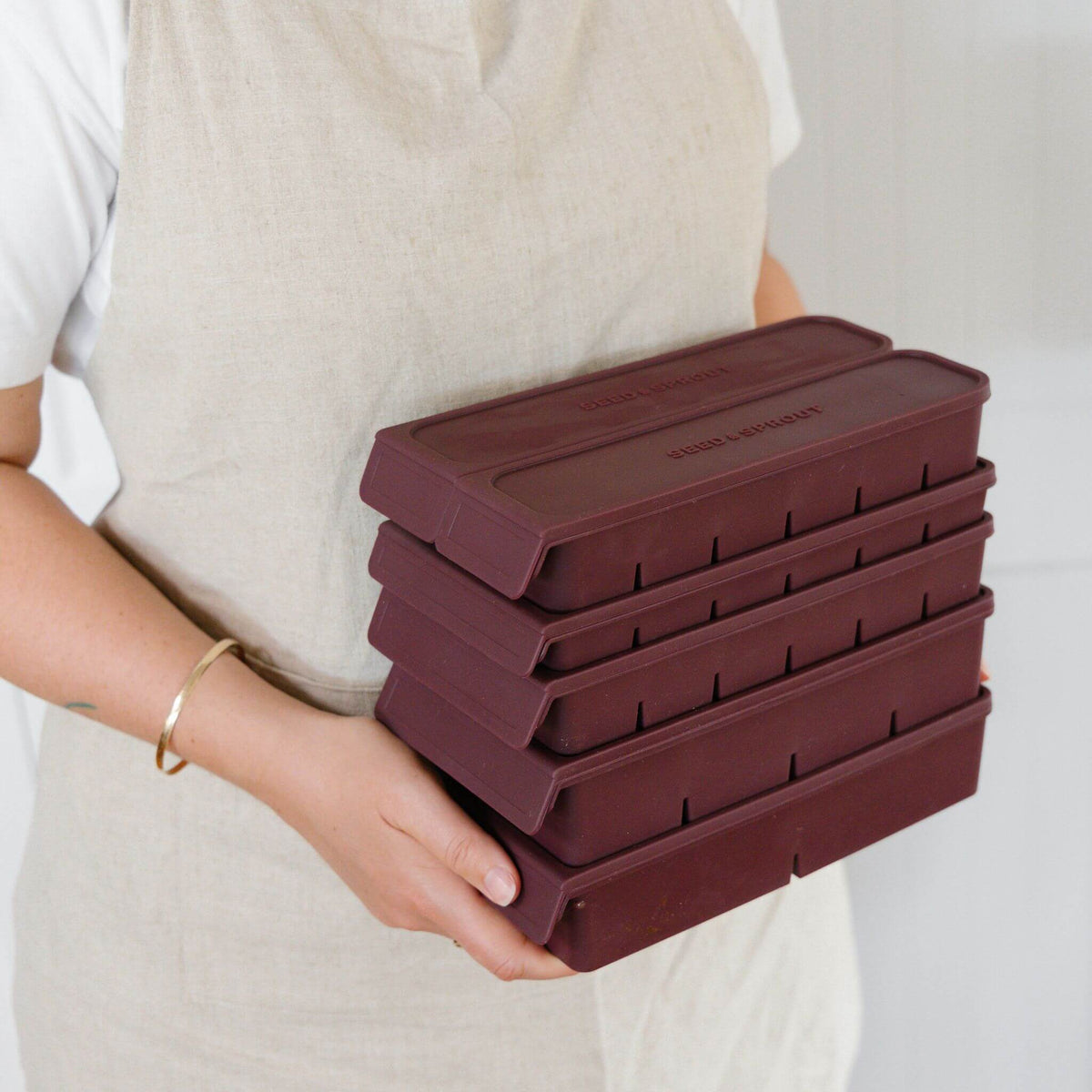 Person in a linen apron holding a full stack of Complete Freezer Tray Set trays in mulberry red, showing the compact stackable profile of the complete set