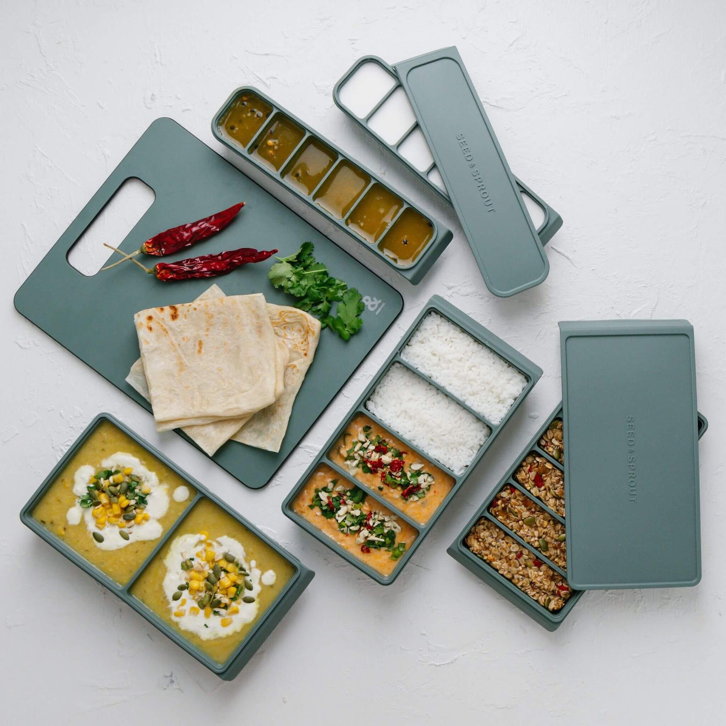 Ultimate Freezer Mould Set in sage green displayed overhead with prepped meals — curry, rice, granola bars, and sauce portions — alongside a matching silicone cutting board with flatbreads, chillies, and fresh herbs